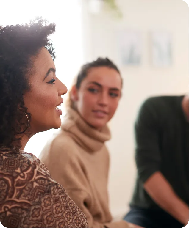 Two women in a room, one with curly hair speaking, another listening attentively.