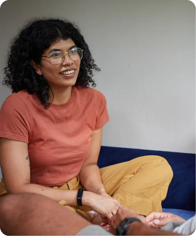 A woman with curly hair and glasses sits on a blue couch, smiling and holding hands with someone off-camera.