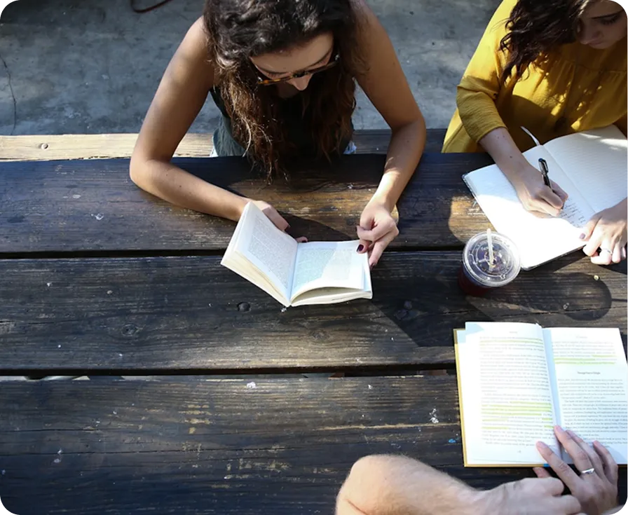 Two people read books at a wooden picnic table, one person's leg is visible in the foreground.