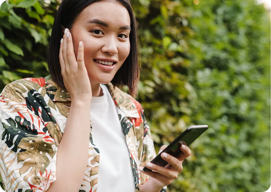 A woman in a floral shirt smiles while holding a phone, with greenery in the background.