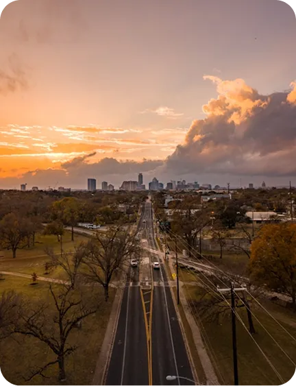 A scenic sunset over a cityscape with a road leading towards the skyline, surrounded by trees and grass.