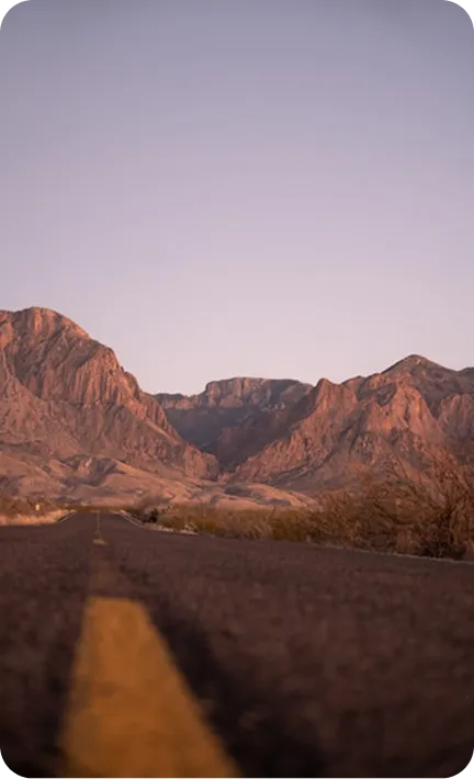A desert road stretches toward rugged mountains under a clear sky.
