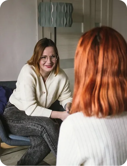 Two women in a cozy room, one with glasses and jeans, the other with red hair, engaged in conversation.
