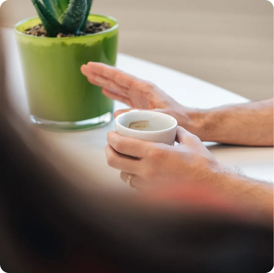 Person holding white cup, hand resting on table, green potted plant in background.