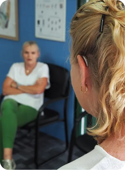 A woman sits with crossed arms in a chair, while another woman listens, wearing a headset.
