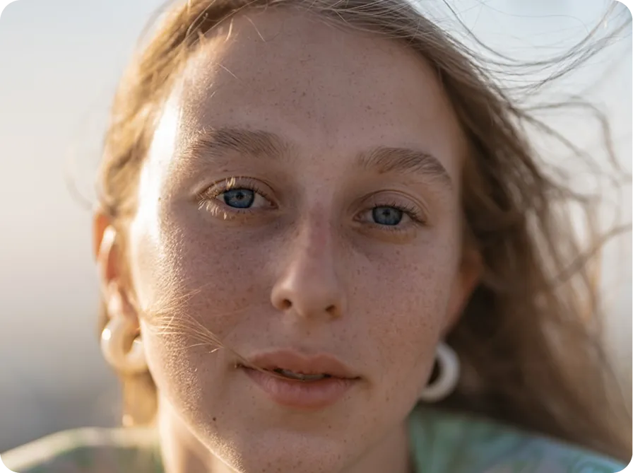 Close-up of a person with freckles, blue eyes, and earrings, hair blowing in the wind.