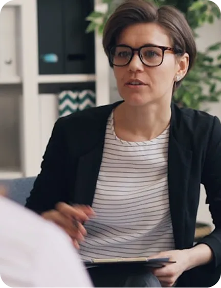 A woman in glasses and a blazer sits in an office, holding a folder, engaged in conversation.