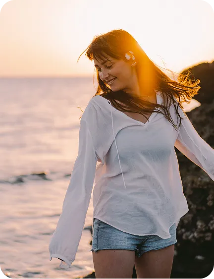A woman in a white shirt and denim shorts smiles near the ocean at sunset.